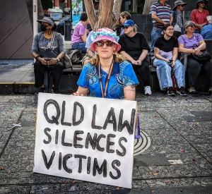 photo shows author TJ Withers at the Brisbane March against domestic violence in April 2024, holding a sign that says Queensland laws silence victims.